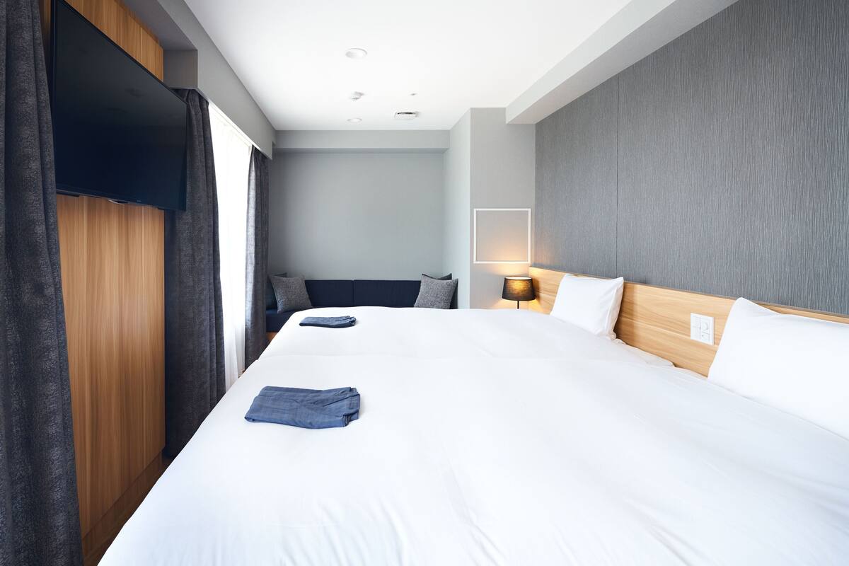 A hotel room with a large bed, a flat-screen TV mounted on the wall, a bedside table with a lamp, and a view of the city through the window.