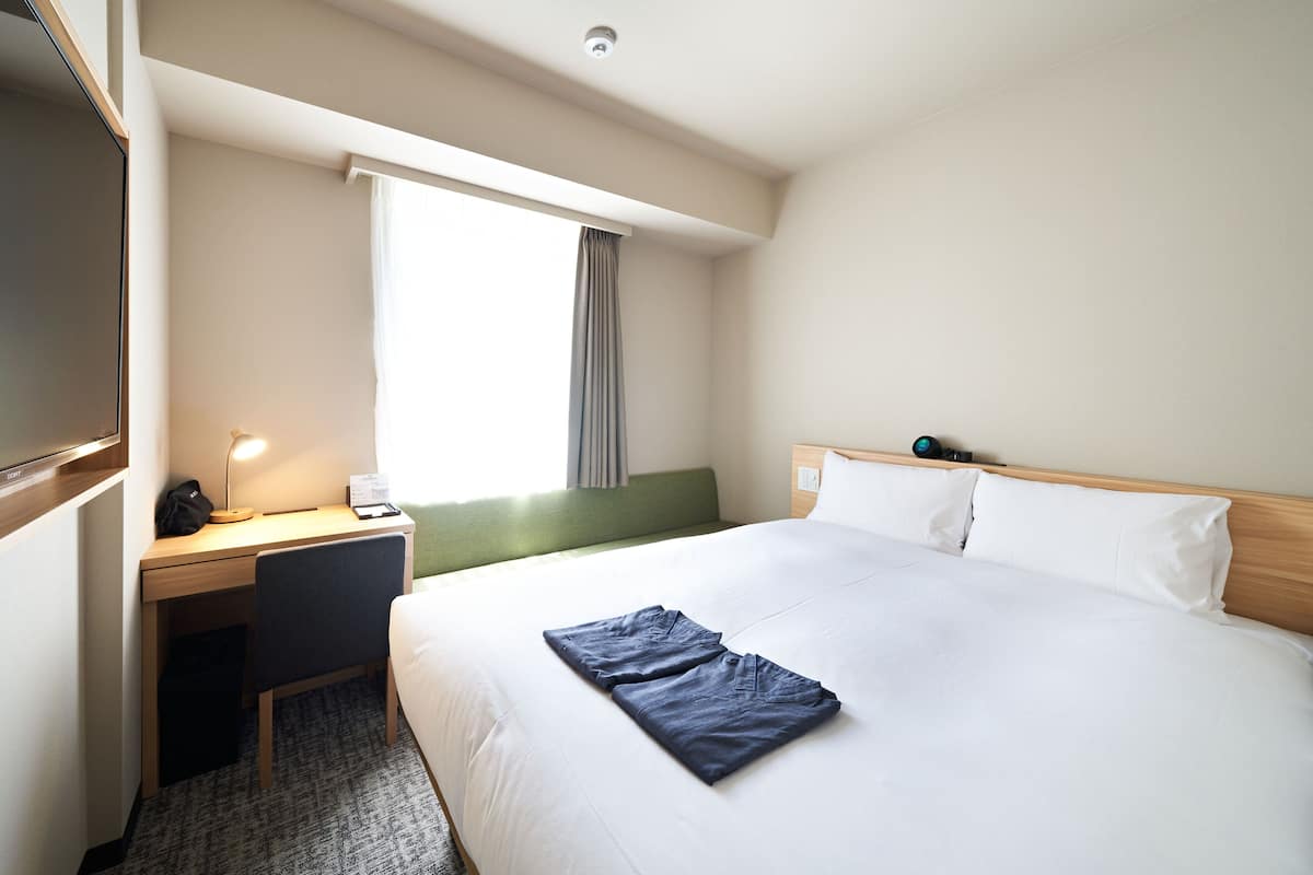 A hotel room with a bed, a desk, a chair, a window with curtains, and a nightstand with a lamp.