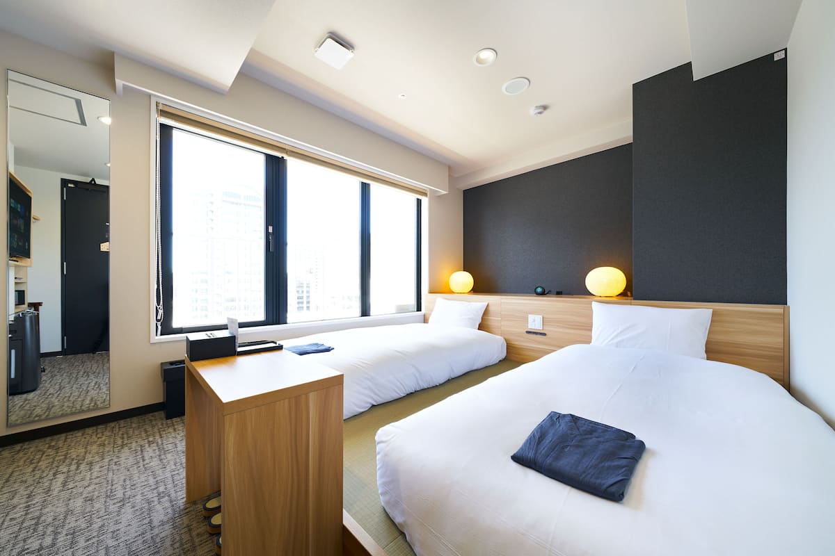 A hotel room with two beds, a desk, a mirror, and a large window.