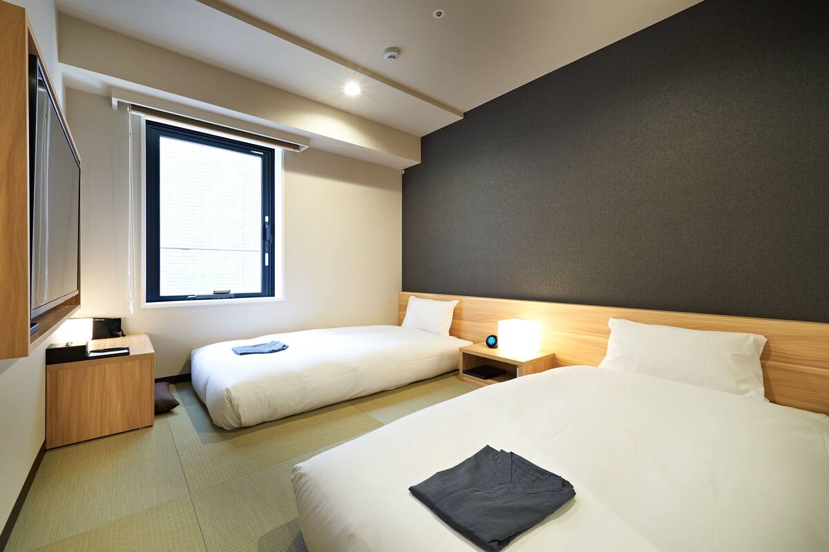 A hotel room with two beds, a bedside table with a lamp, a window with blinds, and a dark wall.