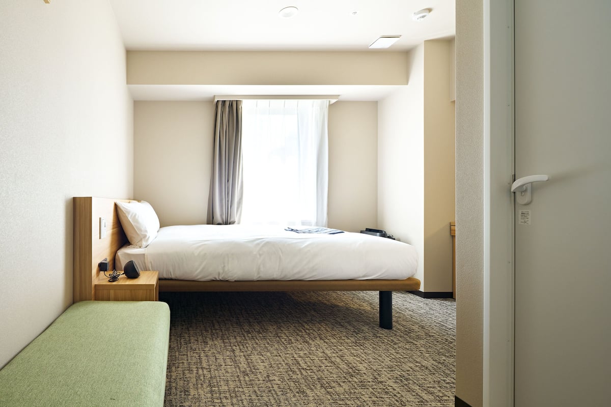 A hotel room with a single bed, a bedside table with a lamp, a green bench, and a window with curtains.