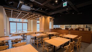 A modern dining area with wooden tables and chairs, a kitchen in the background, and large windows allowing natural light. - HOTEL Enmichi (Kawasaki)