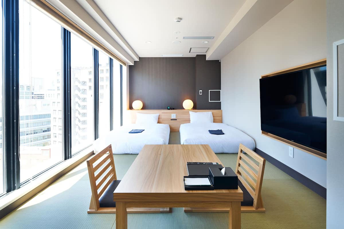 A modern hotel room with a large window, two beds, a wooden table, and a flat-screen TV.