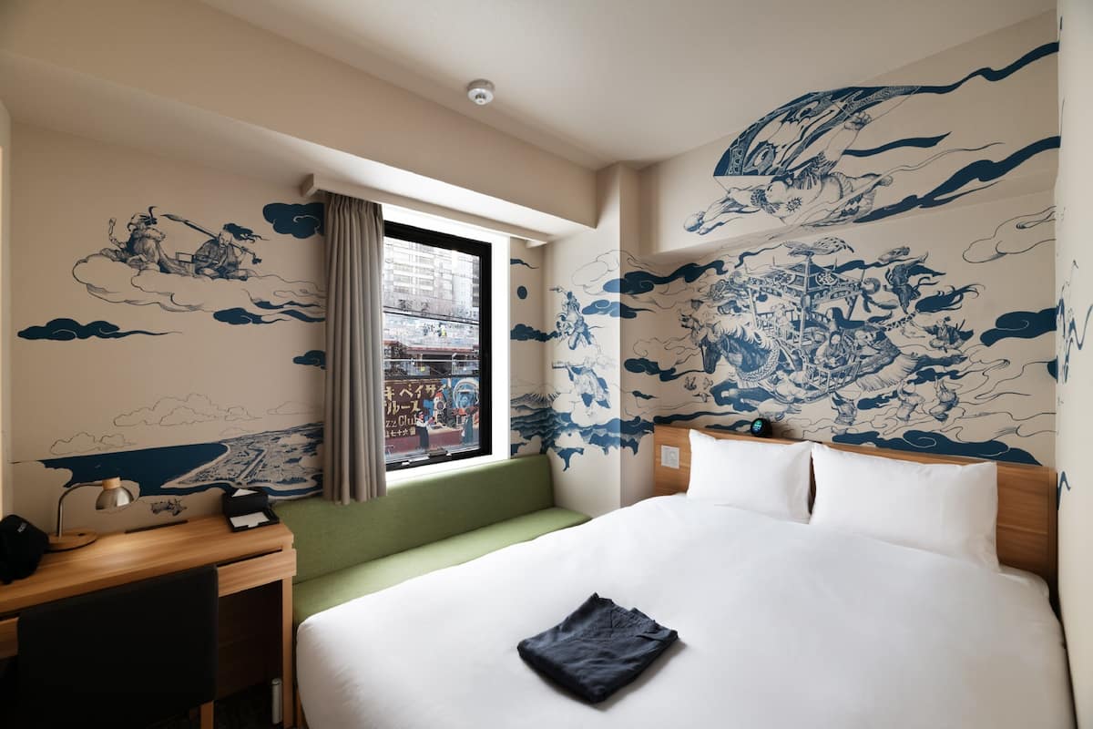A hotel room with a large bed, a desk with a lamp, a window with curtains, and traditional wall art.