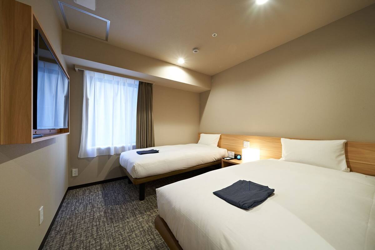 A hotel room with two beds, a TV, a window with curtains, a nightstand with a lamp, and a carpeted floor.
