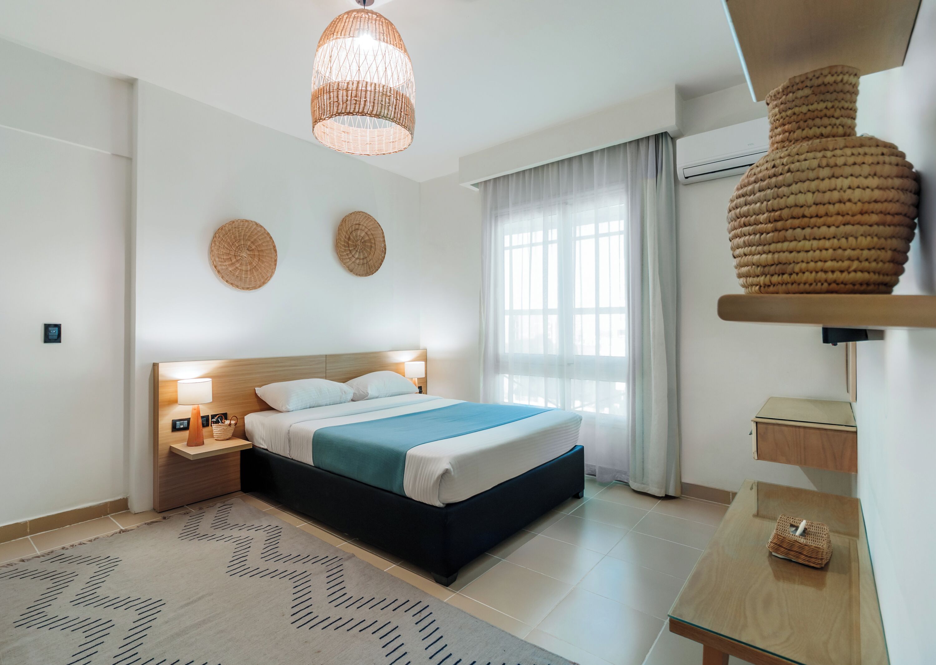 A modern hotel room with a large bed, bedside lamps, a woven light fixture, a large window with sheer curtains, and a small table with a basket.