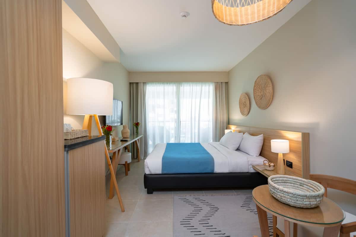 A hotel room with a bed, bedside table, lamp, and a small round table with a woven basket.