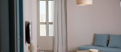 A room with a sofa, a round table, a hanging light fixture, a window with curtains, and a television on the wall.