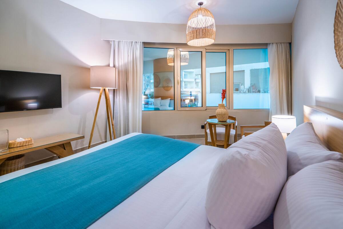 A hotel room with a large bed, a flat-screen TV, a small table, and a window with curtains.