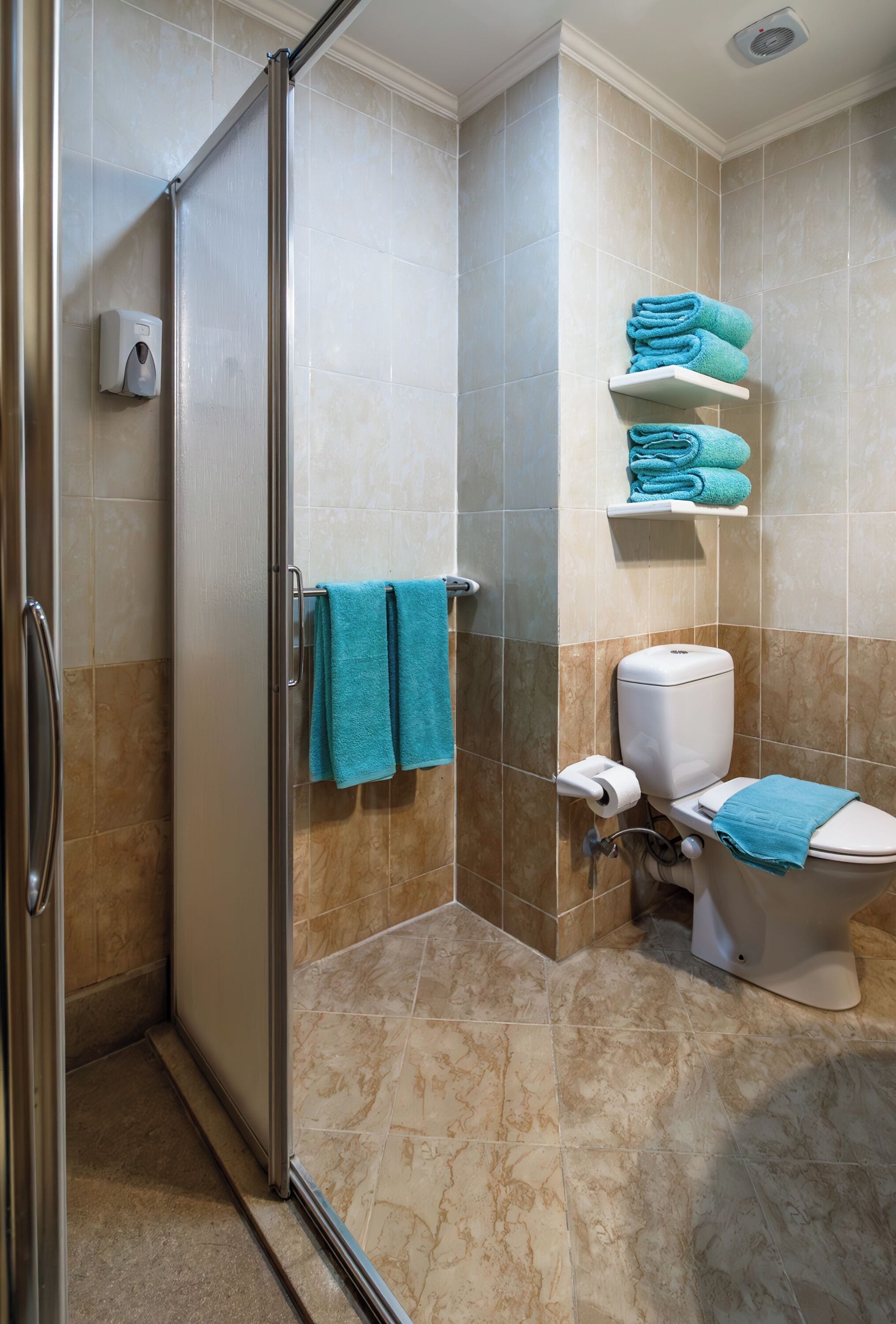 Un baño con mampara de vidrio, un inodoro con una toalla azul y un estante con toallas dobladas.