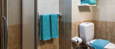 Un baño con mampara de vidrio, un inodoro con una toalla azul y un estante con toallas dobladas.