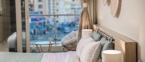 Ein Hotelzimmer mit Bett, Kissen, einer Nachttischlampe und Blick auf die Stadt durch große Fenster.