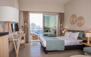 Habitación de hotel con cama, escritorio, silla, balcón con vistas a la ciudad y dos obras de arte en las paredes.