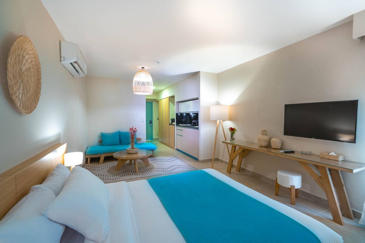A modern hotel room with a large bed, a desk with a flat-screen TV, a seating area with a sofa and a small table, and a kitchenette in the background.