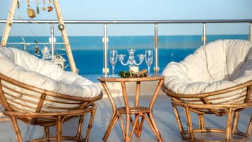 Two wicker chairs with cushions, a small round table with a candle and glasses, and a view of the sea through a balcony railing.