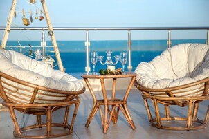 Dos sillones de mimbre con cojines, una mesita redonda con una vela y vasos, y vista al mar a través de la barandilla de un balcón.