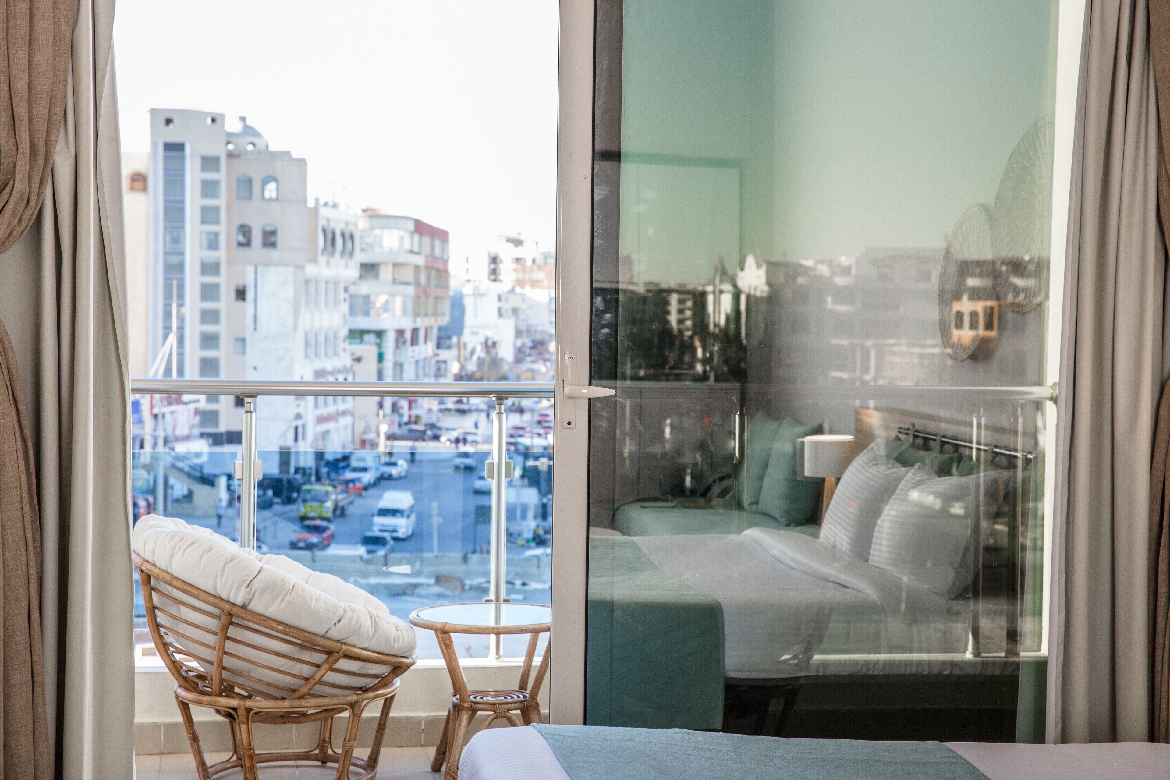 Un balcón con una silla de mimbre y una mesita, una cama con edredón verde, y vista a la ciudad a través de un ventanal.