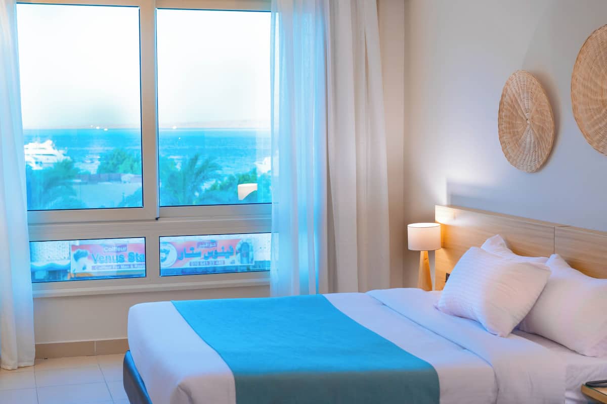A hotel room with a large window offering a view of the sea and buildings, a bed with white bedding, and a wall-mounted lamp.