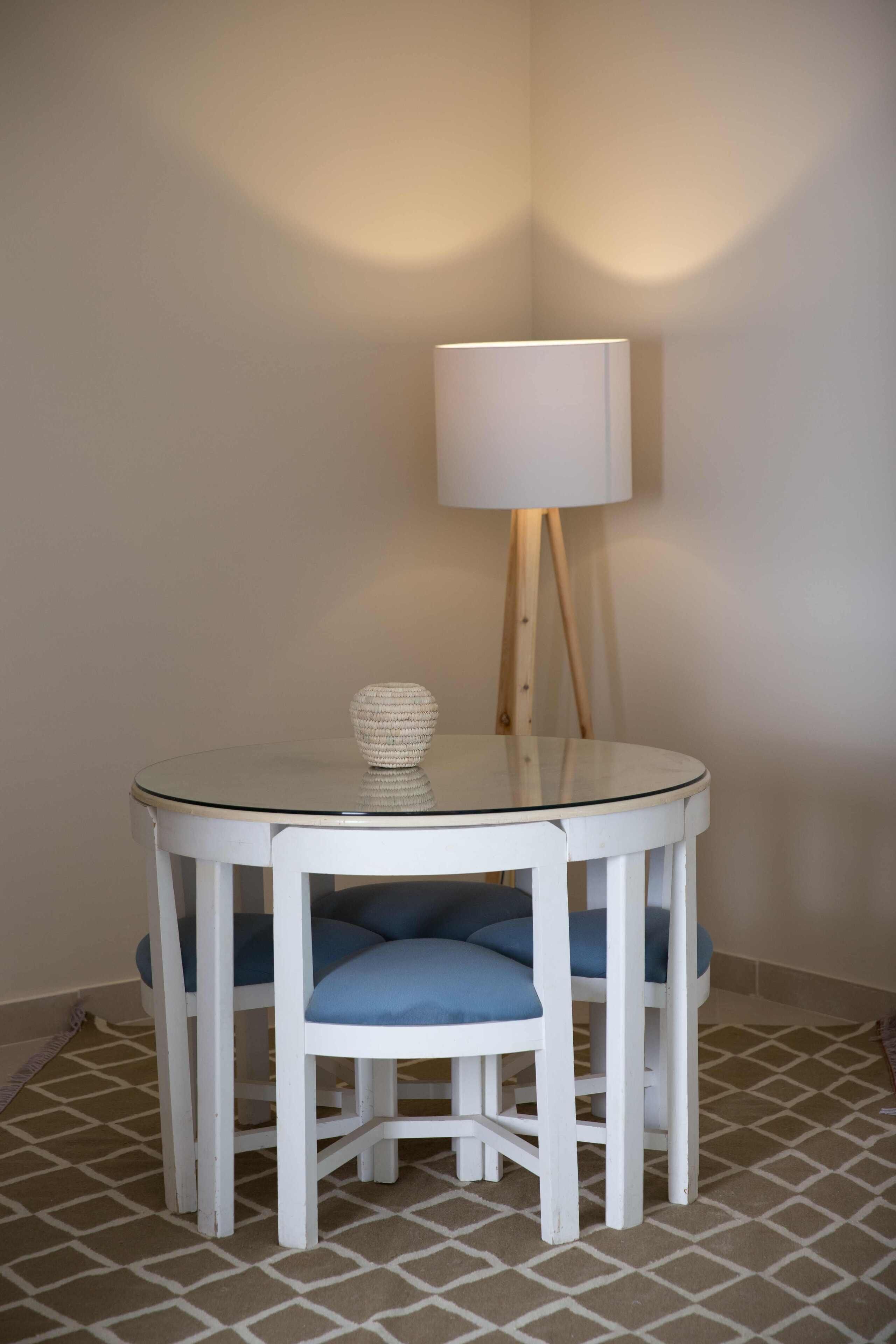 A round glass table with white legs and blue cushioned chairs, a white lamp with a wooden stand, and a patterned floor.