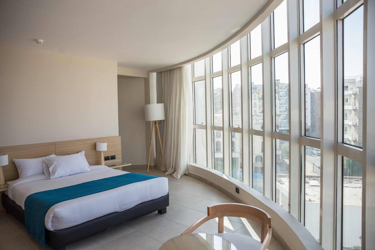 A hotel room with a large bed, a chair, and a view of the city through large windows.