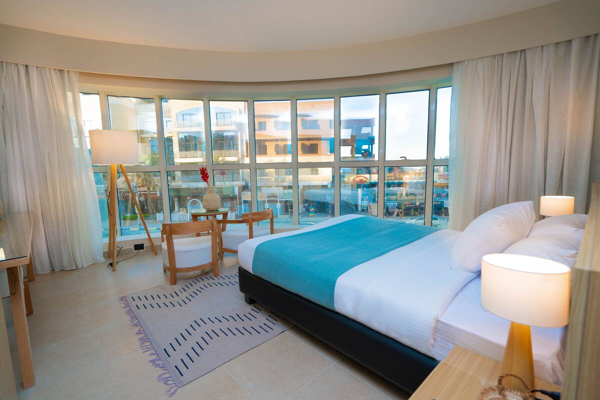 A hotel room with a large bed, a desk, and a view of the city through large windows.