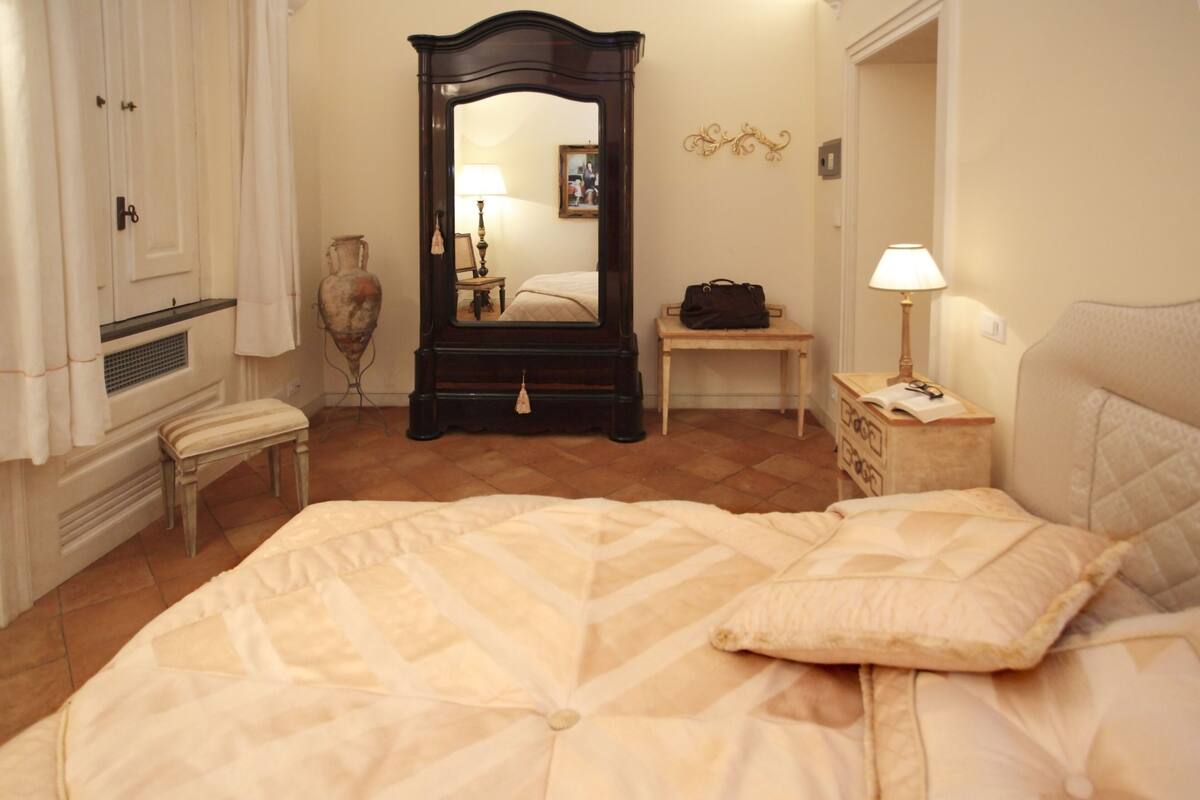 Une chambre à coucher comprenant un lit, une commode, une coiffeuse avec un miroir, un banc, une lampe et un tableau accroché au mur.