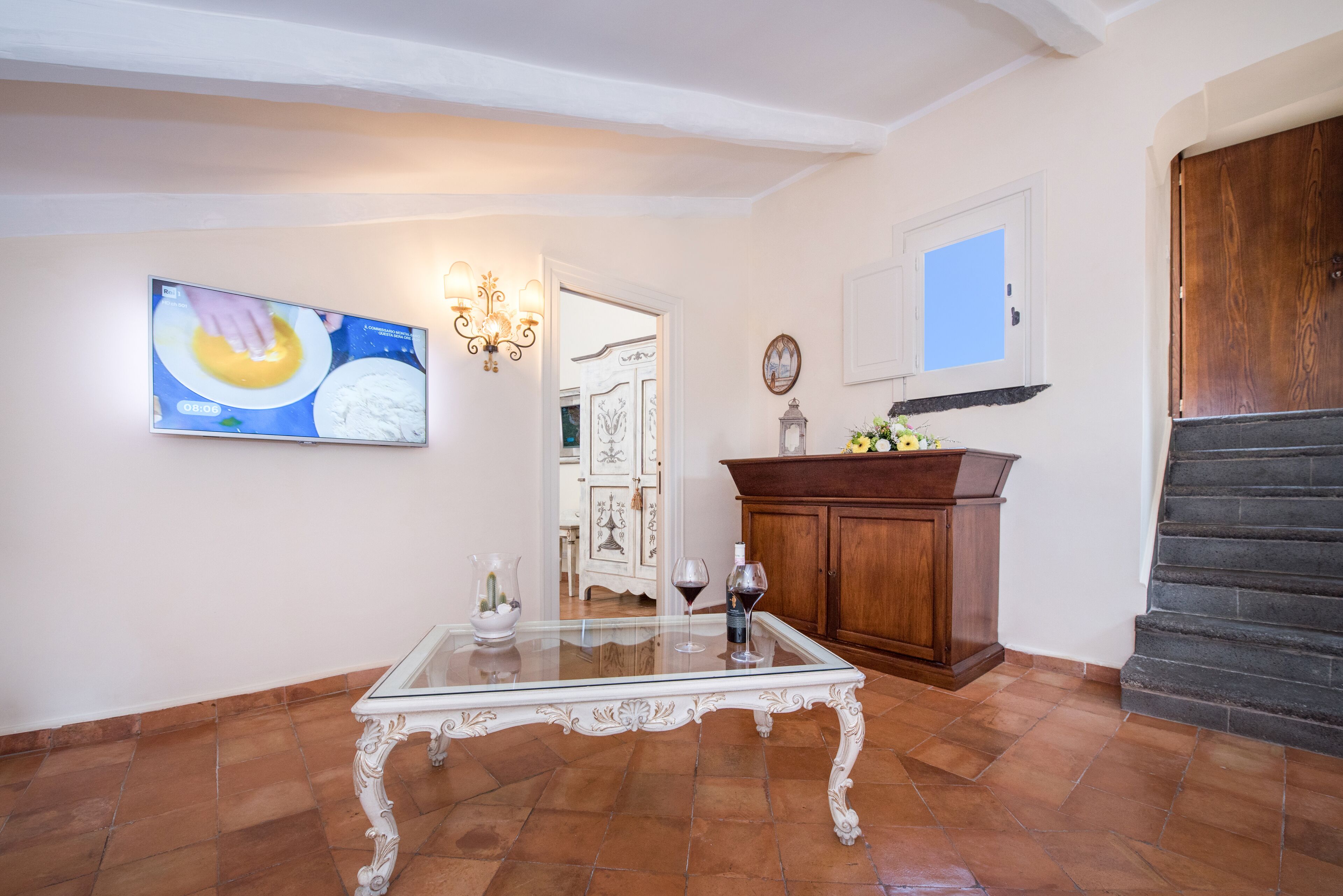 A spacious living room with a glass coffee table, a wooden cabinet, and a television displaying a cooking program.
