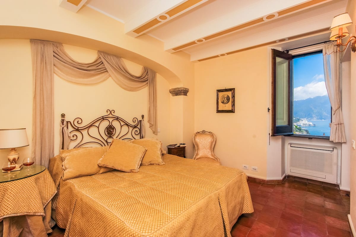 Une chambre à coucher avec un lit, une table de chevet, une lampe, une fenêtre donnant sur un beau paysage et un tableau au mur.