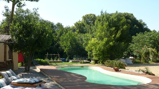 A swimming pool with a wooden deck, surrounded by gravel, trees, and outdoor furniture. - Superb Gîte in Mas (Bollène)