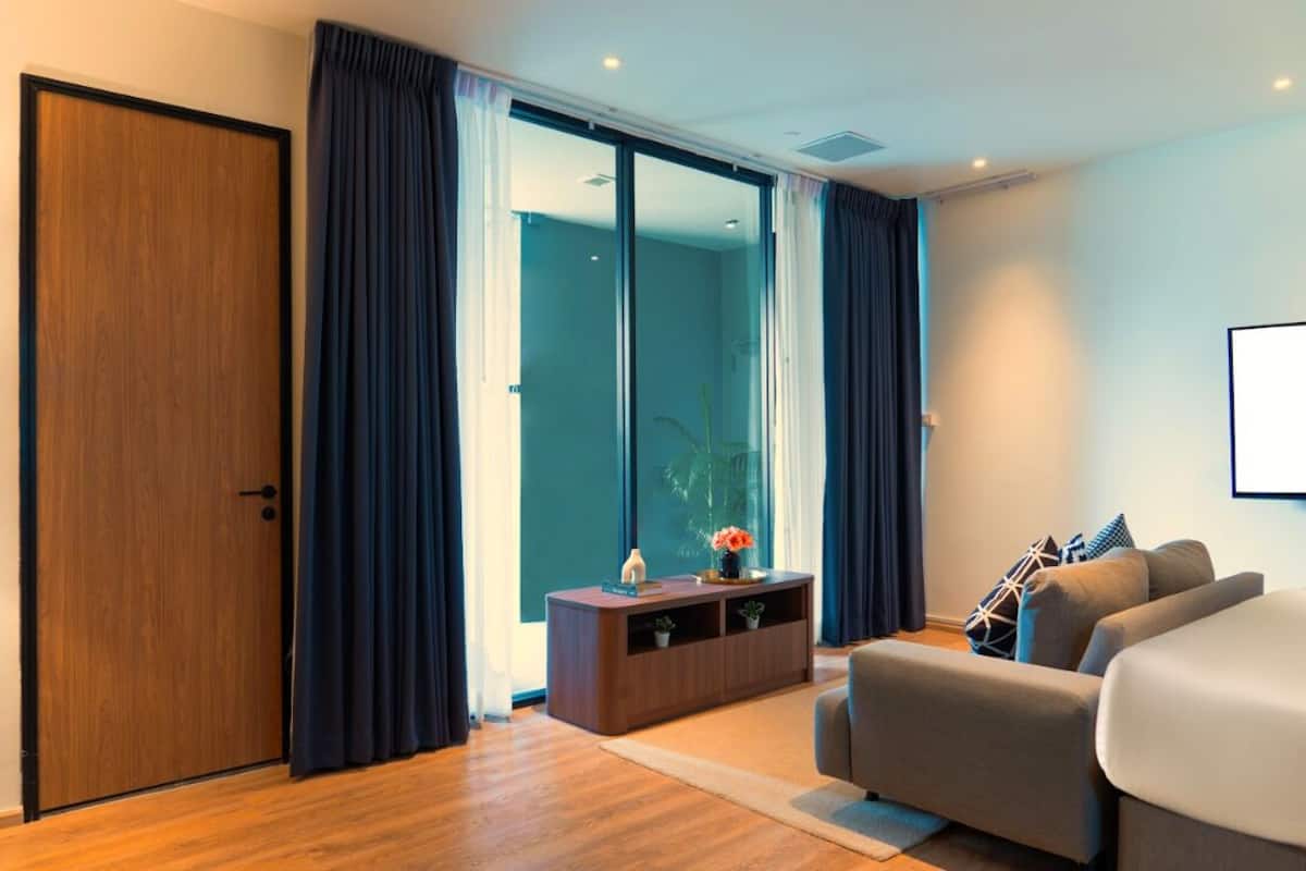 Une chambre d’hôtel moderne avec une porte en bois, des rideaux foncés, une grande fenêtre, un canapé, un petit meuble et un lit.
