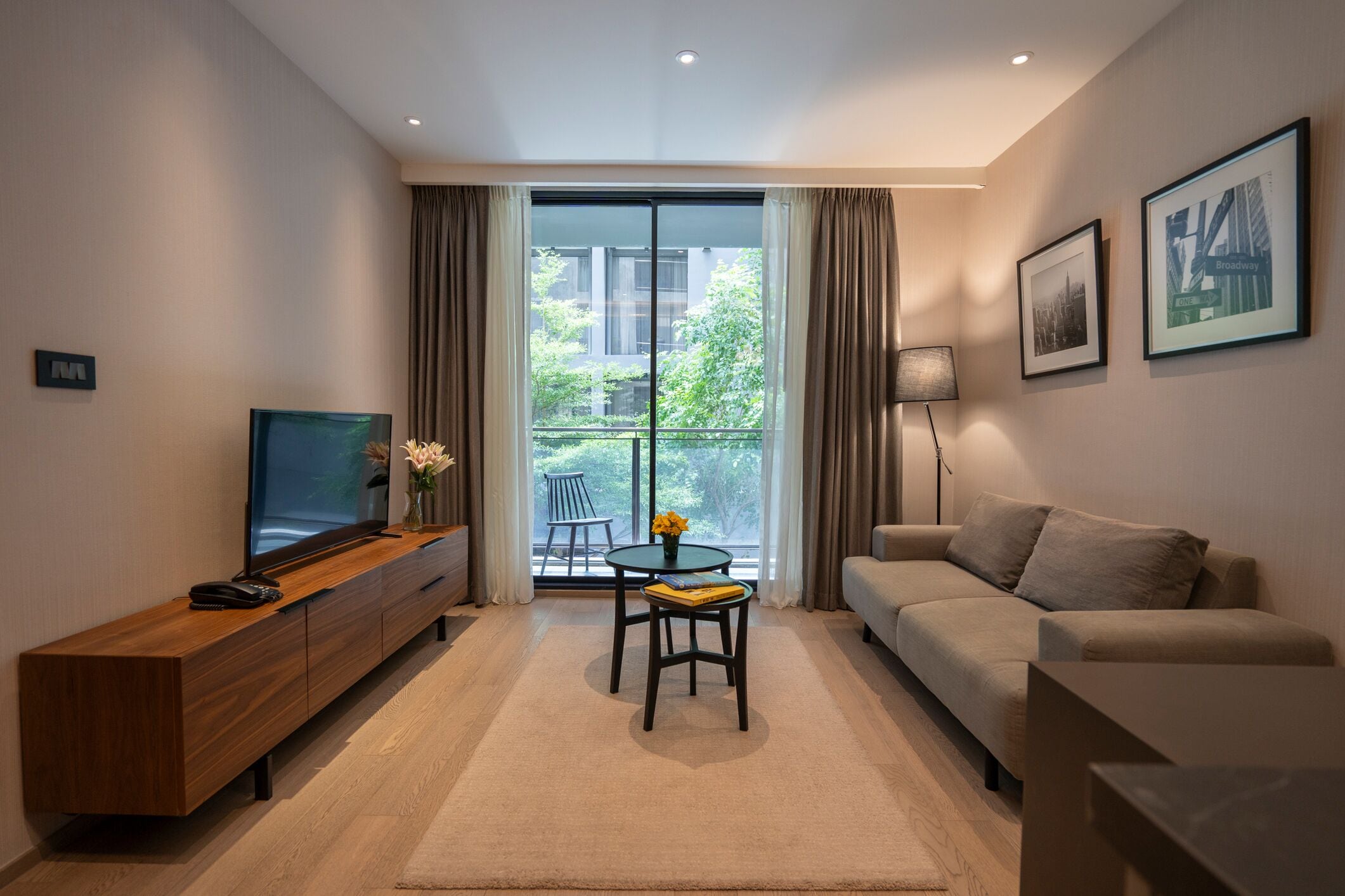 A modern living room with a sofa, a TV on a wooden console, a small table with a vase, and a large window with curtains.
