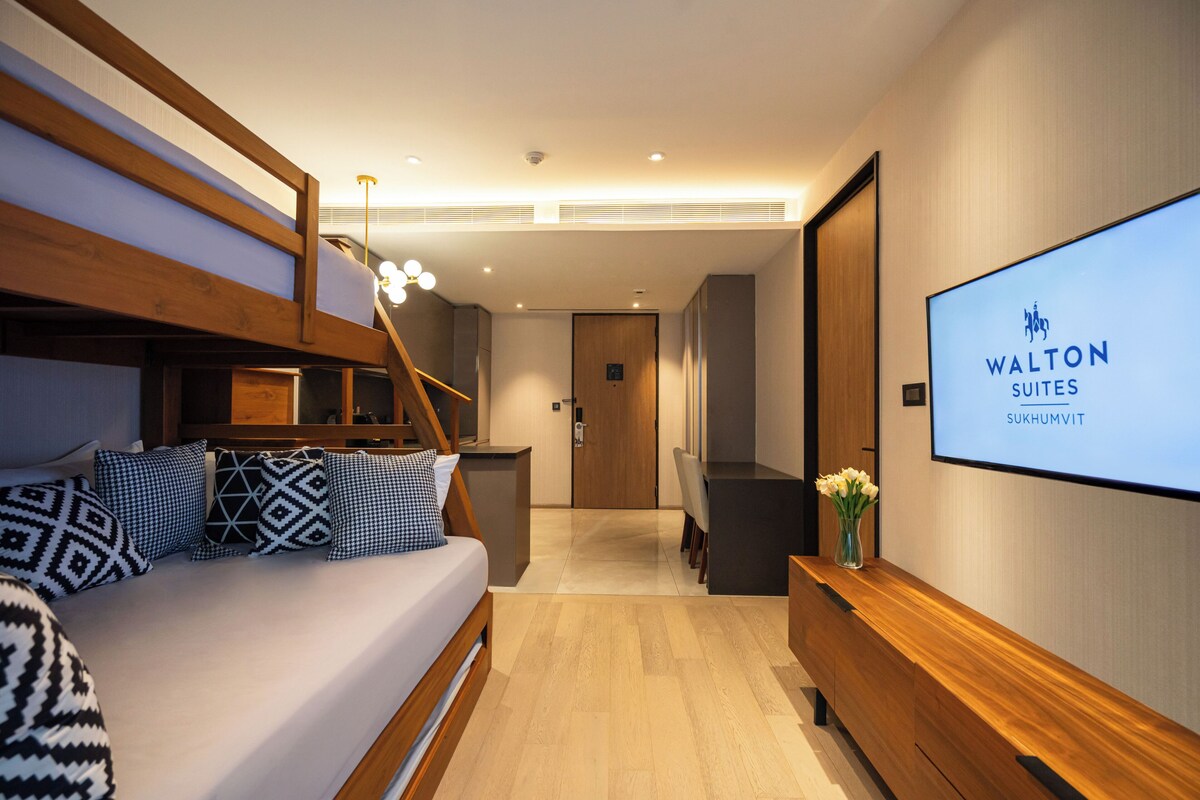 Une chambre d’hôtel avec un lit superposé, un bureau, un écran de télévision affichant le nom de l’hôtel et un banc en bois.