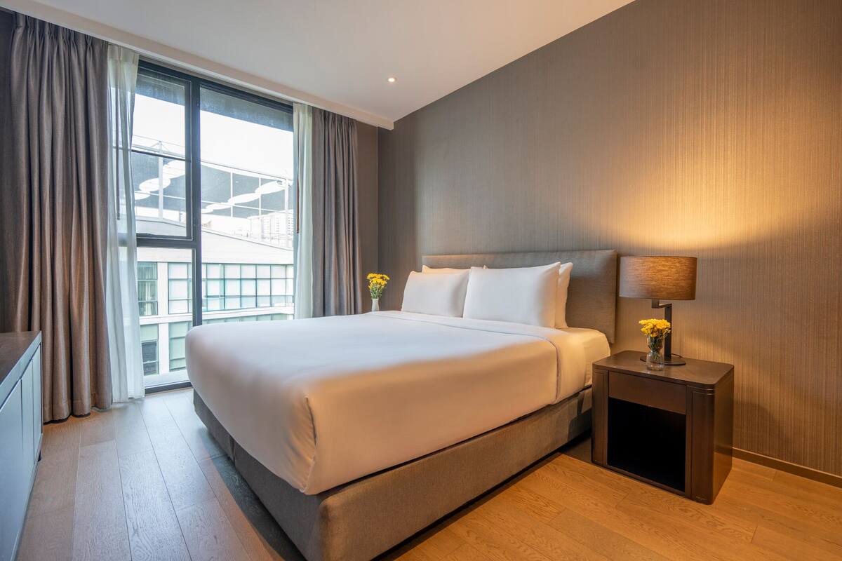 Une chambre d’hôtel avec un grand lit, une table de chevet avec une lampe et une vue sur un immeuble par la fenêtre.
