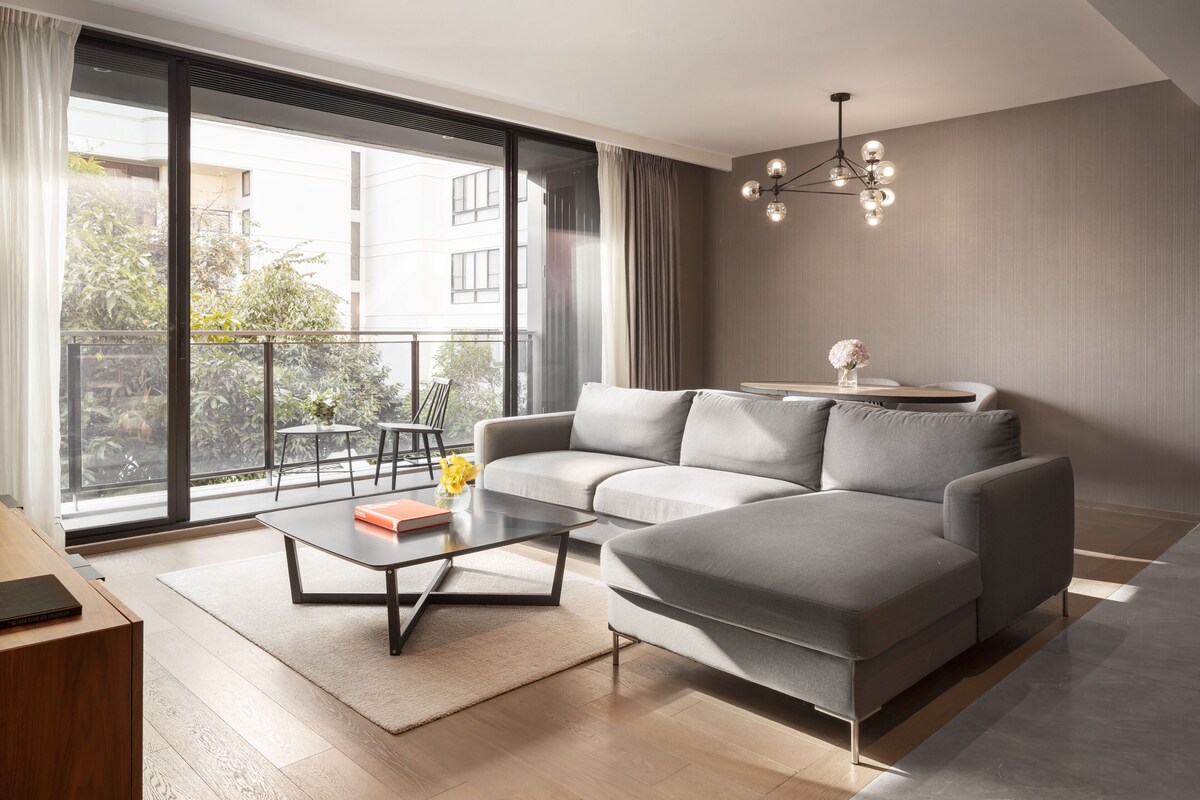 Un salon moderne avec un canapé d’angle gris, une table basse en verre et une vue sur un balcon agrémenté de plantes, avec un immeuble en arrière-plan.