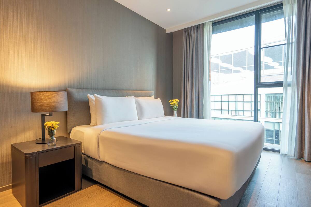 Une chambre d’hôtel moderne avec un grand lit, une table de chevet avec une lampe et une vue sur la ville par la fenêtre.