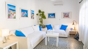 A modern living room with two white sofas, a small white table, and a potted plant. There are framed pictures on the wall and a lamp on a side table. - Rena Hotel (Santorini)
