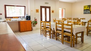 A dining area with wooden chairs and tables, a coffee station, and a potted plant. - Rena Hotel (Santorini)