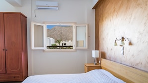 A bedroom with a bed, a wooden wardrobe, a nightstand with a lamp, a window with an open view of buildings and a mountain, and an air conditioning unit. - Rena Hotel (Santorini)