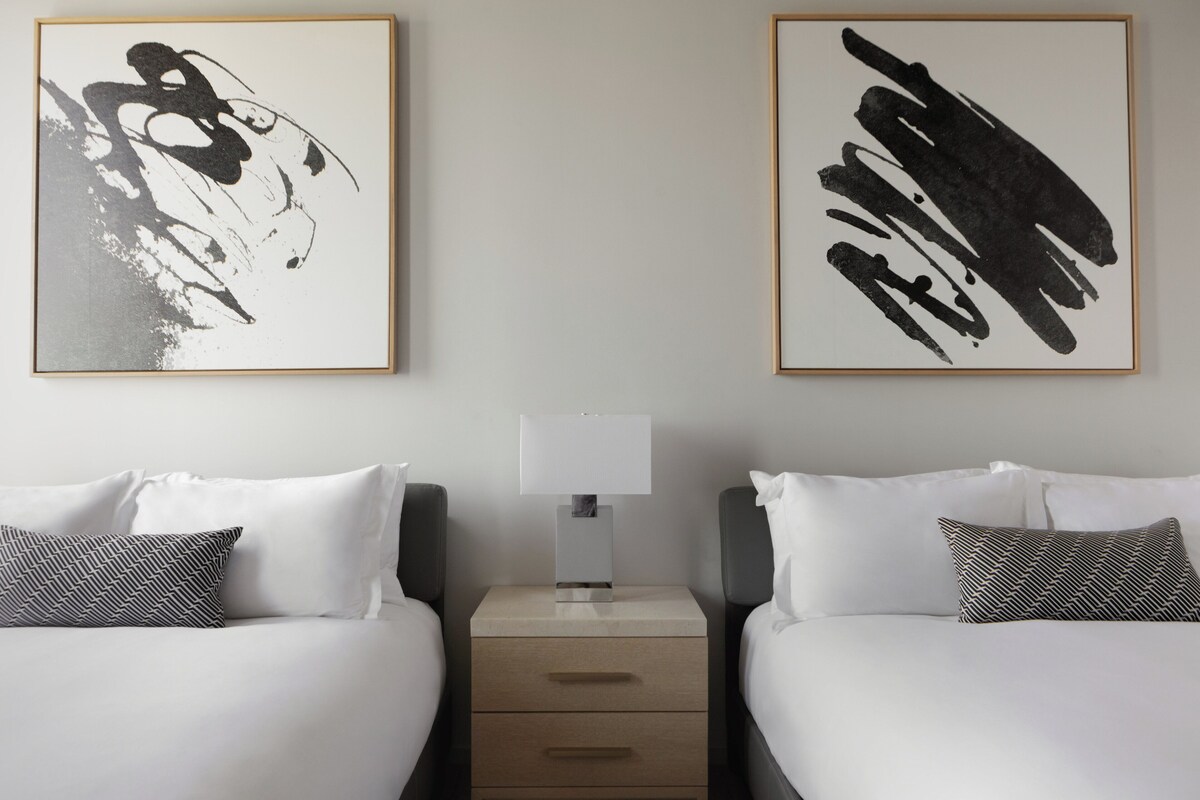 A hotel room with two beds, a nightstand with a lamp, and two framed abstract paintings on the wall.