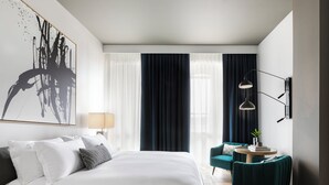 A modern hotel room with a large bed, a desk with a chair, a large abstract painting, and a view of the city through the window.