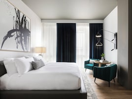 A modern hotel room with a large bed, a desk with a chair, a large abstract painting, and a view of the city through the window.