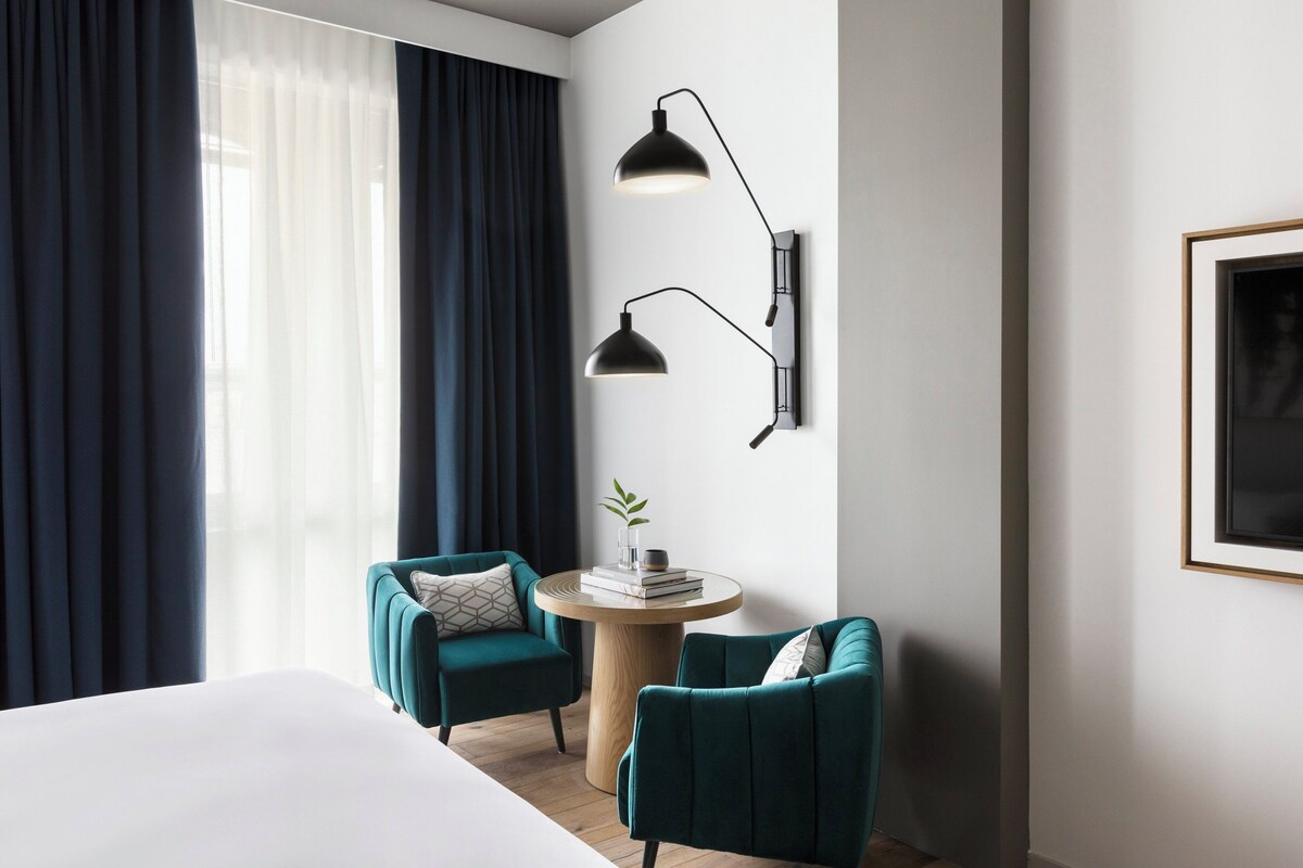 A hotel room with a bed, two teal armchairs, a round wooden table with books, a wall-mounted lamp, and a framed picture.