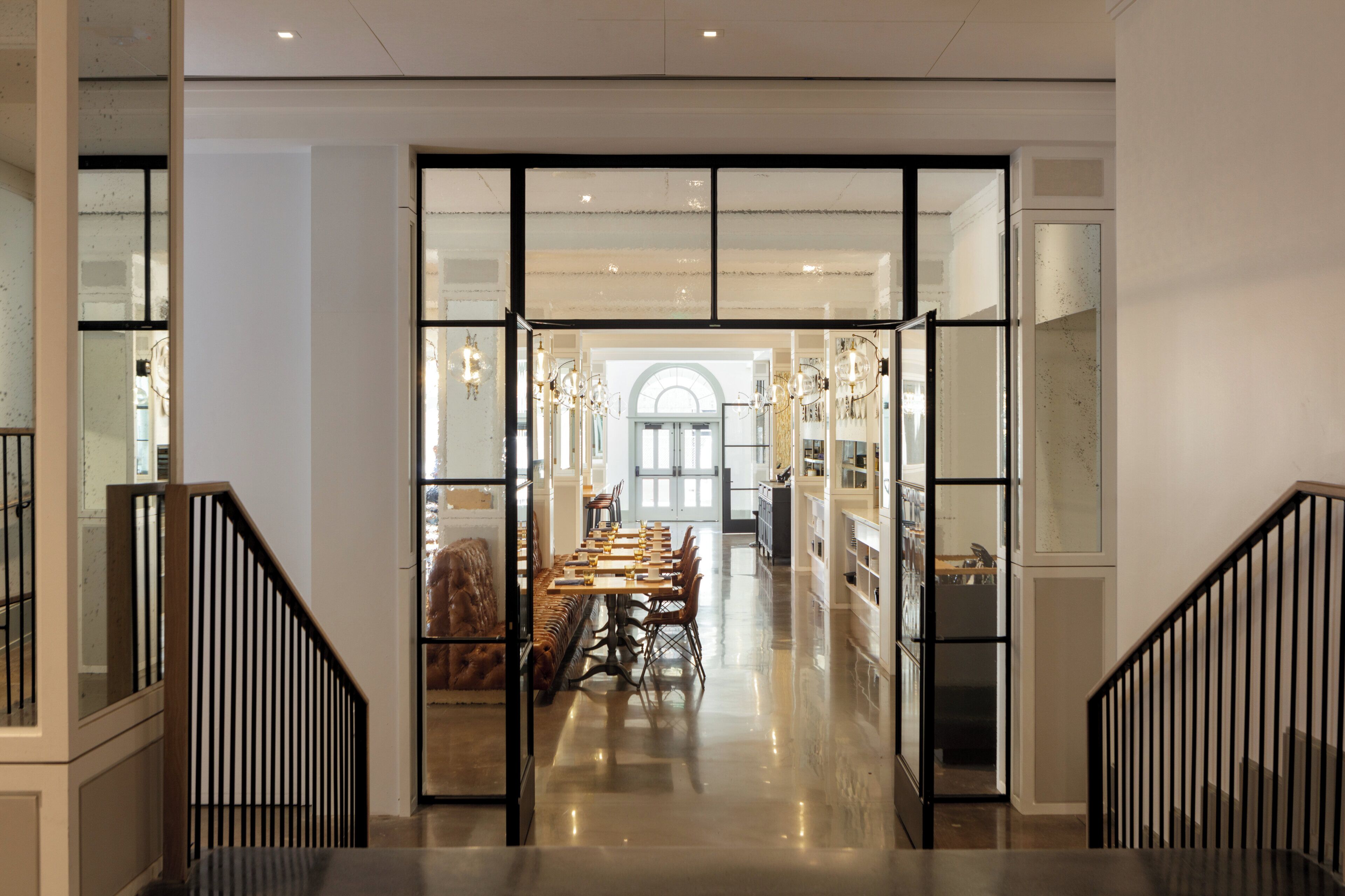 Un intérieur spacieux comprenant un coin repas central, un escalier et une vue sur une autre pièce dotée d’une grande fenêtre.