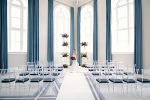 A wedding ceremony setup with a bride in a white dress, clear chairs, and blue curtains.