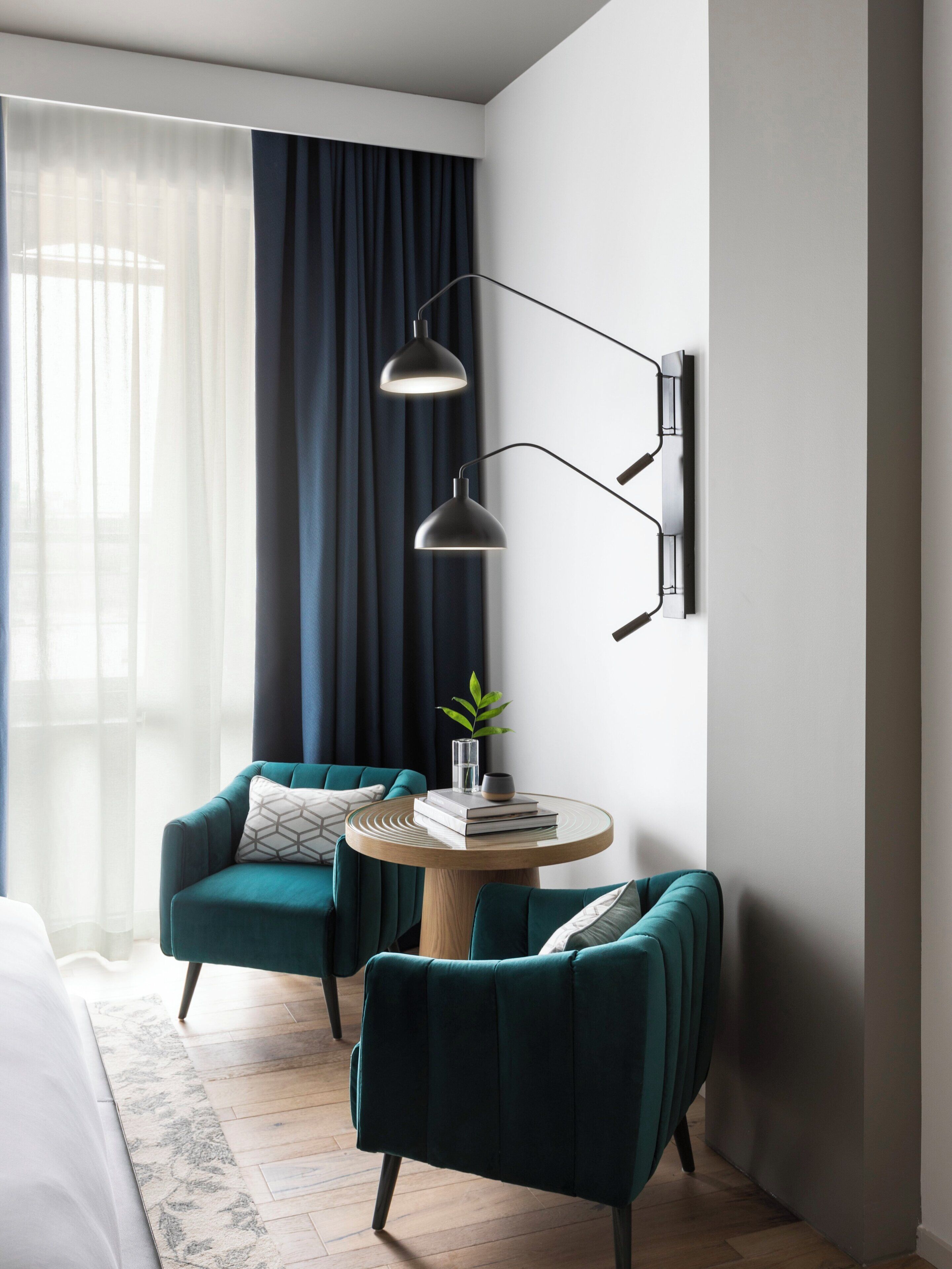 A modern room with two teal armchairs, a round wooden table with books and a plant, and a wall-mounted lamp.