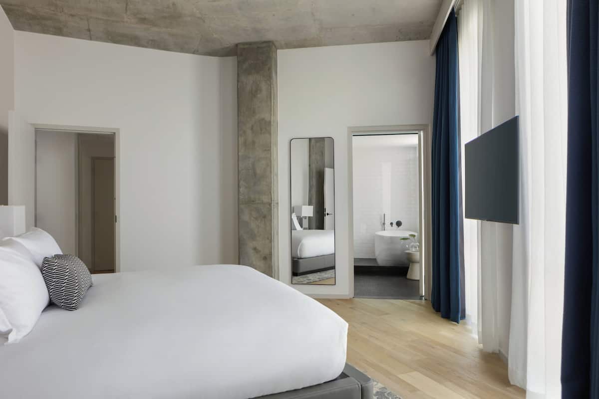 A modern hotel room with a large bed, a flat-screen TV, a mirror, and a bathroom visible through an open door.