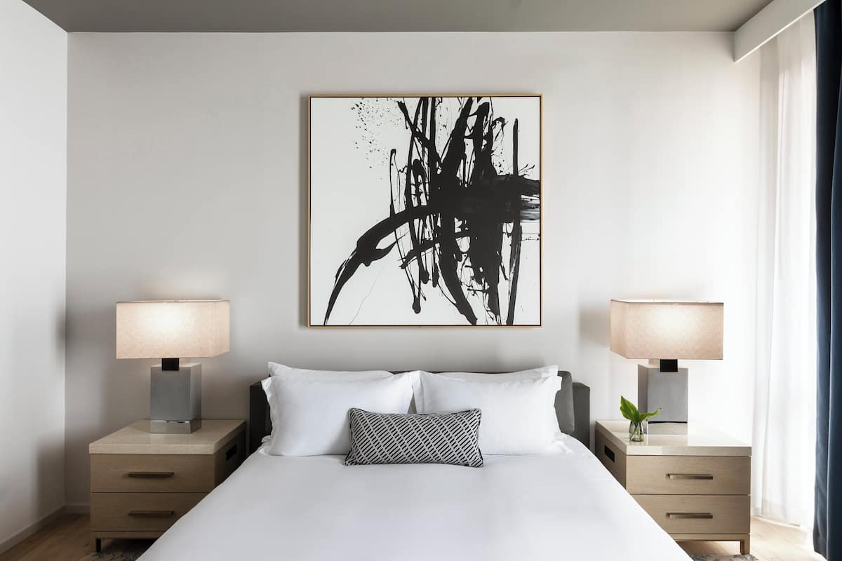 A modern bedroom with a large bed, two bedside tables with lamps, a black and white abstract painting, and a small plant on one table.