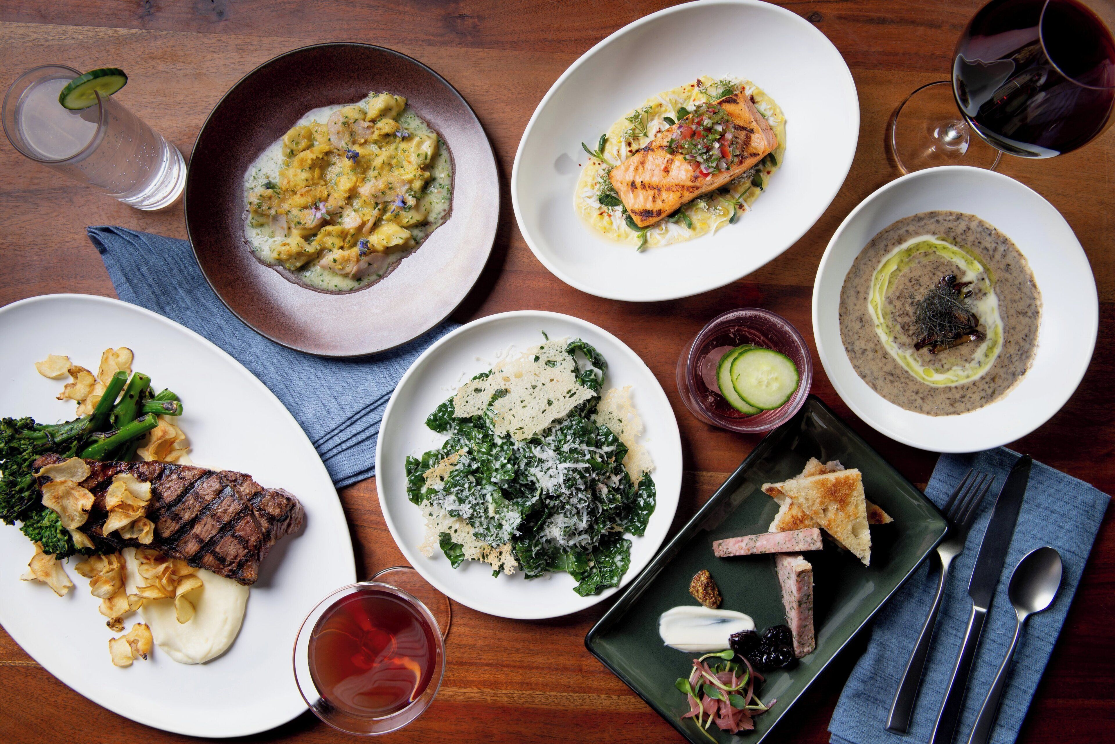 Variedad de platos sobre una mesa de madera, incluyendo un bistec con champiñones, un plato de verduras y un tazón de sopa.