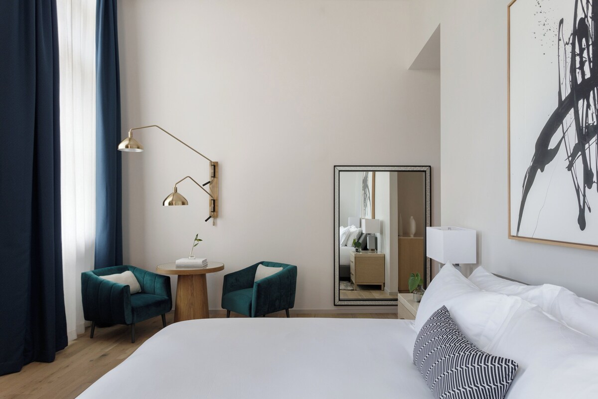 A hotel room with a large bed, two chairs, a small table, a lamp, a mirror, and a painting on the wall.