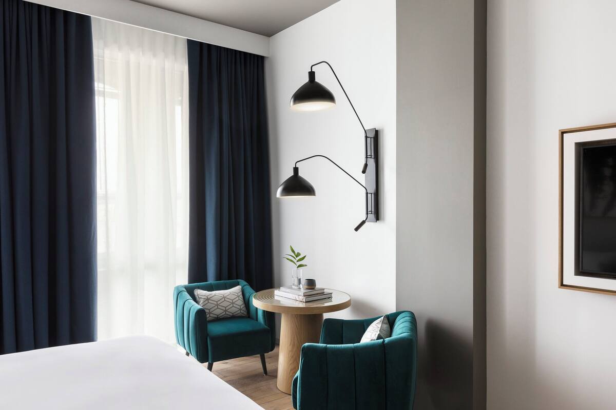 A modern hotel room with a large bed, two teal armchairs, a small round table with books, a wall-mounted lamp, and a framed picture on the wall.